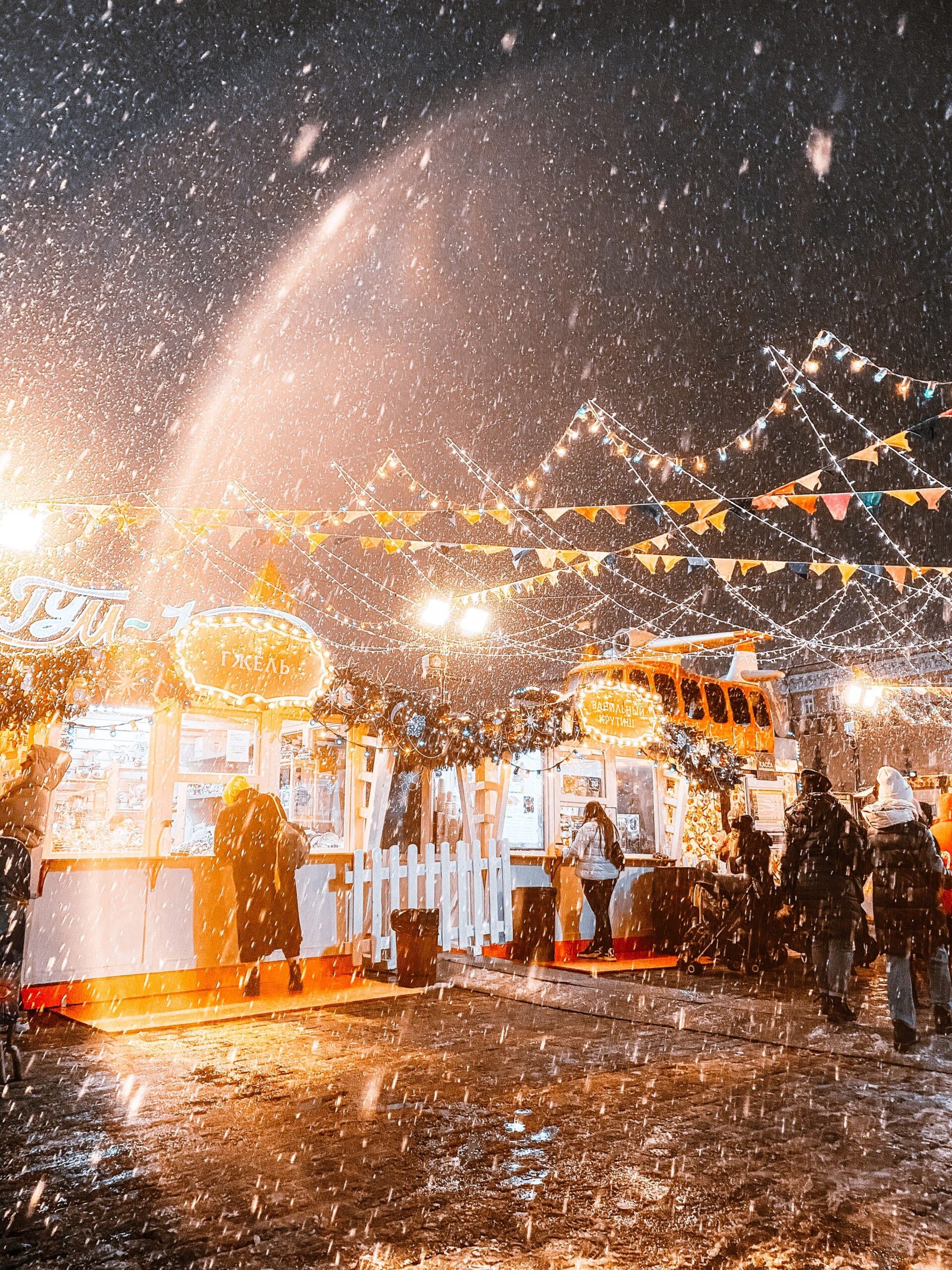 Winter street market lit by string lights under falling snow at night
